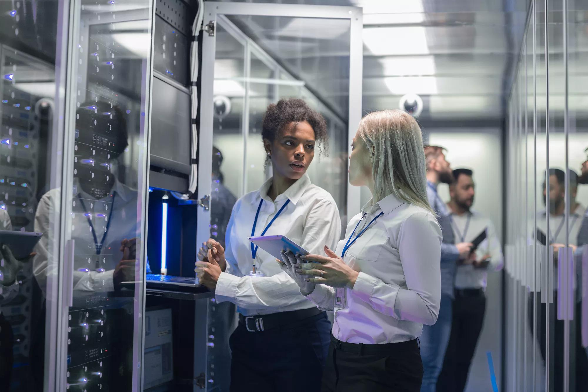 Medium shot of two women working in a data center with rows of server racks and checking the equipment and discussing their work