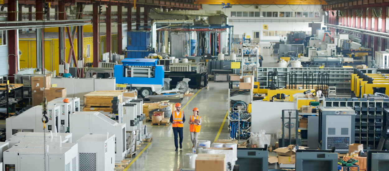 a man and a woman, both in orange vests and orange hard hats walk side-by-side through a warehouse