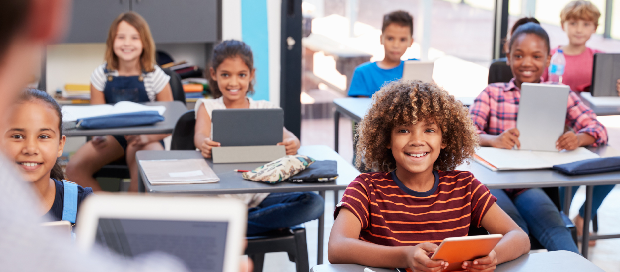 young children with smiles on their faces sit at their desks with tablets in front of them