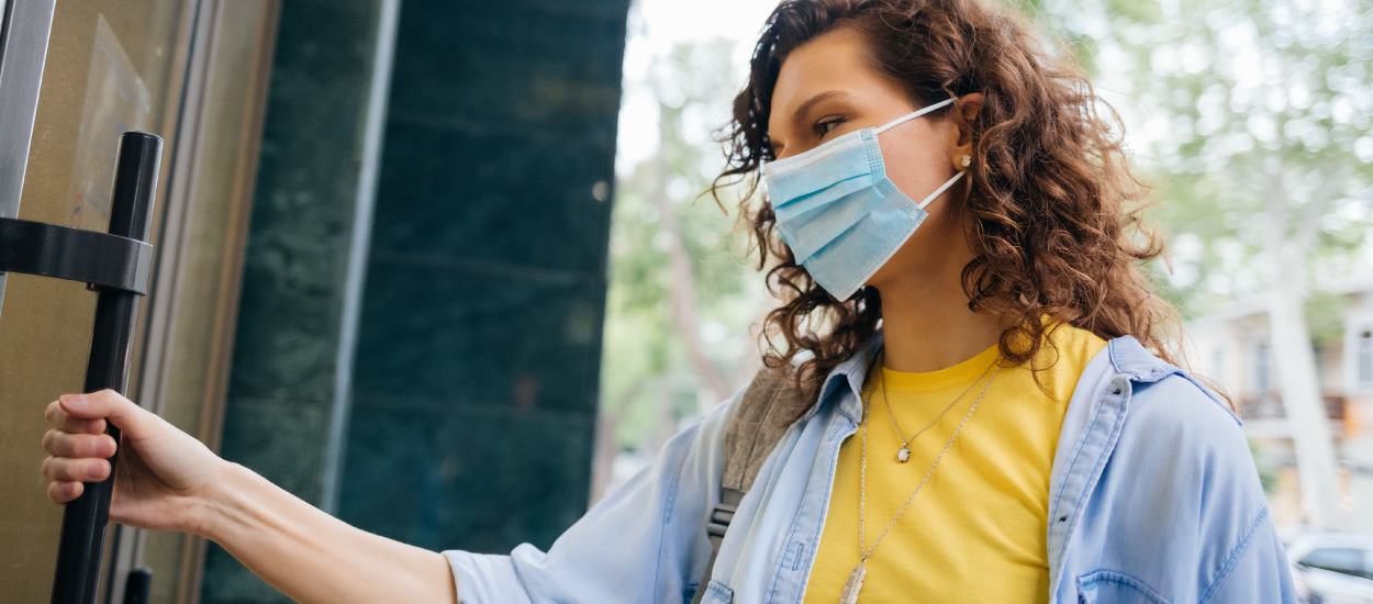 a woman wearing a yellow shirt and light blue jacket, with a mask on her face, is opening a door