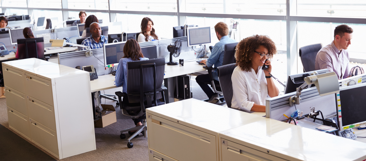 People in business attire sit in an open space workplace, talking on phones and working on their computers.