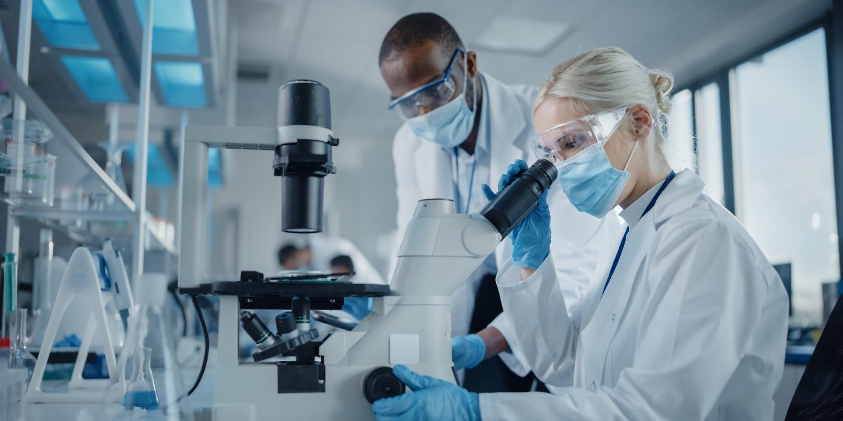 Modern Medical Research Laboratory: Two Scientists Wearing Face Masks Working Together Using Microscope, Analysing Samples, Talking. Advanced Scientific Lab for Medicine, Biotechnology.