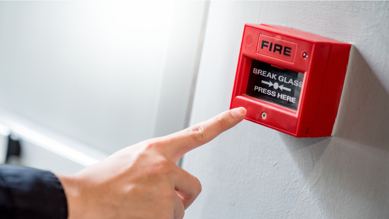 Male hand pointing at red fire alarm switch on concrete wall in office building