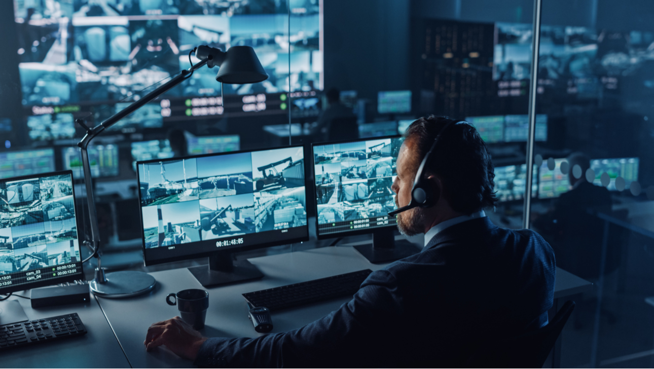 Male Officer Works on a Computer with Surveillance CCTV Video in a Harbour Monitoring Center with Multiple Cameras on a Big Digital Screen