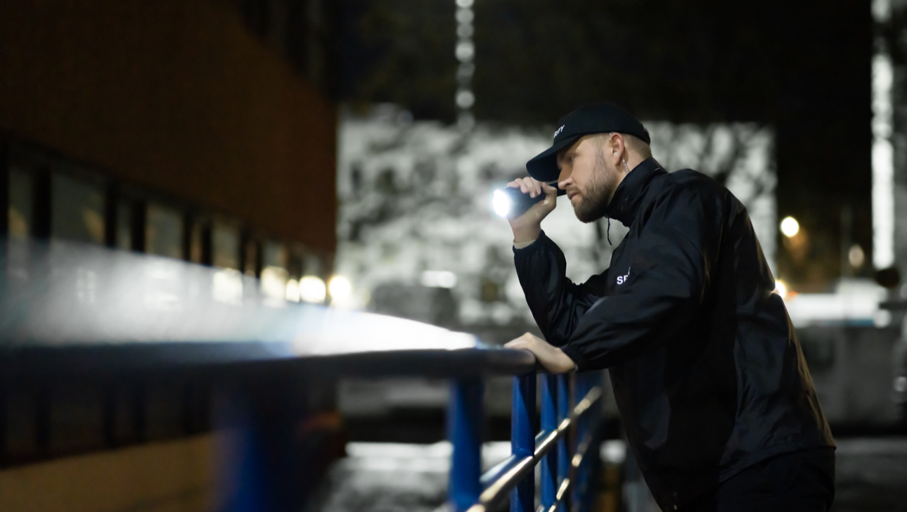 Security Guard Walking Building Perimeter With Flashlight At Night