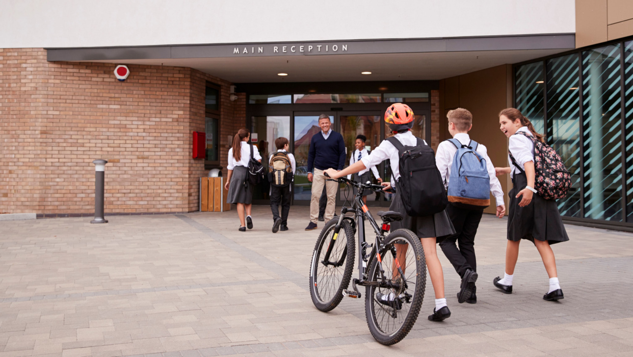 Group Of High School Students Wearing Uniform Arriving At School Walking Or Riding Bikes Being Greeted By Teacher