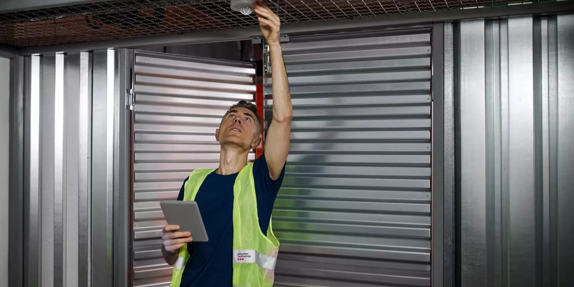 Fire alarm. Serious focused responsible man with tablet checking fire safety adjusting device on garage ceiling