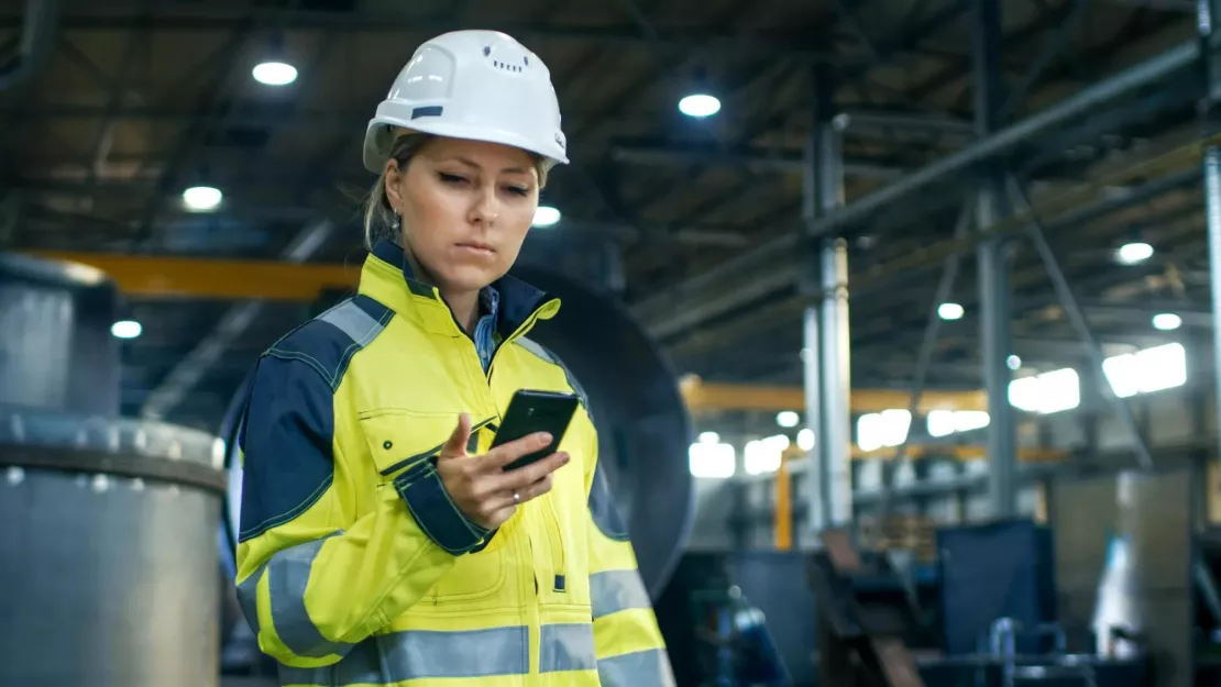 Female Industrial Worker in the Hard Hat Uses Mobile Phone While Walking Through Heavy Industry Manufacturing Factory. In the Background Various Metalwork Project Parts Lying