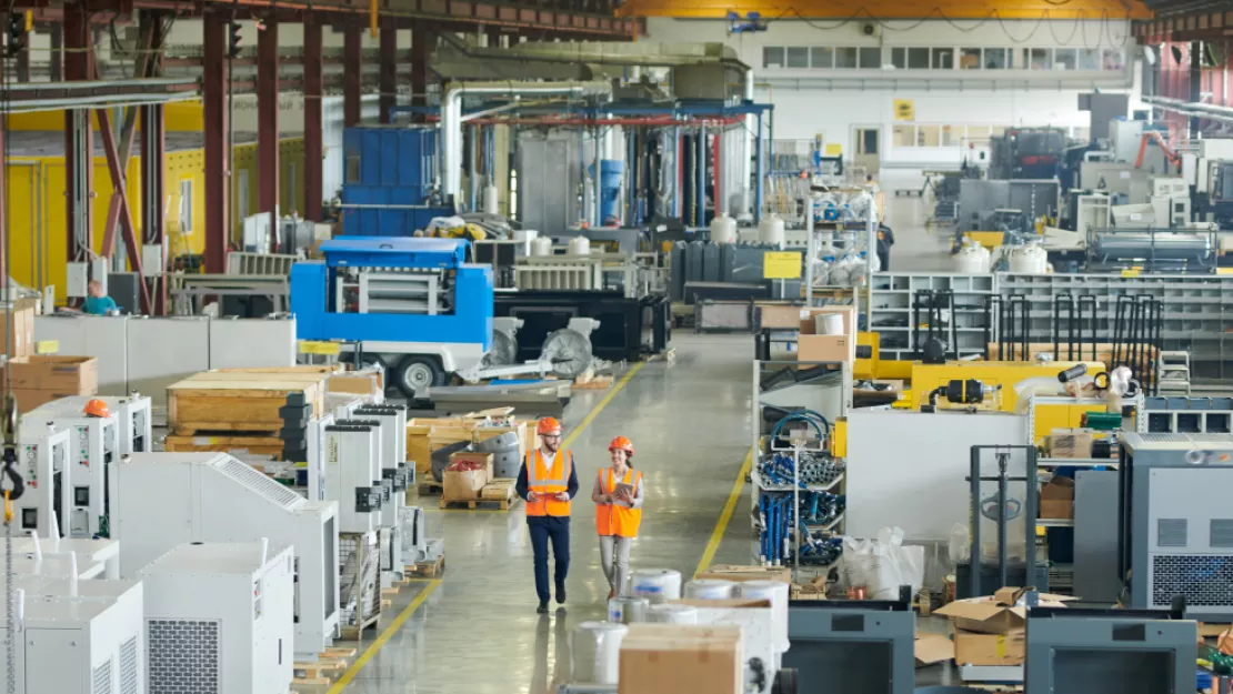 a man and a woman, both in orange vests and orange hard hats walk side-by-side through a warehouse