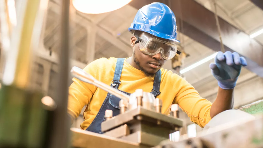 A man in a yellow shirt and blue hard hat, goggles, and blue gloves works on machinery