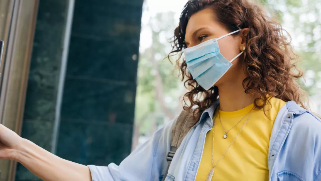 a woman wearing a yellow shirt and light blue jacket, with a mask on her face, is opening a door