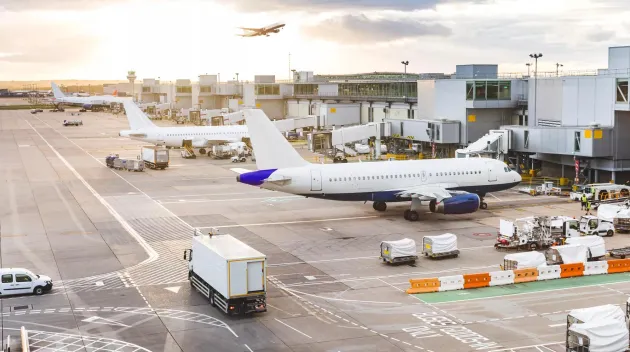 Busy airport view with airplanes and service vehicles at sunset. London airport with aircrafts at gates and taking off, trucks all around and sun setting on background. Travel and industry concepts