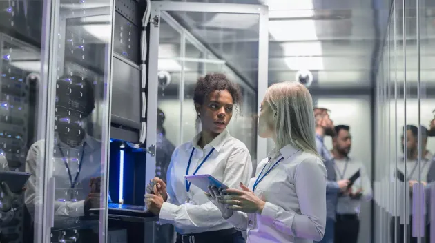 Medium shot of two women working in a data center with rows of server racks and checking the equipment and discussing their work