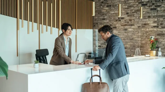 Traveling During COVID-19: Businessman with Luggage Passing Passport and Talking with Concierge on a Hotel Reception with Sneeze Guard Protection