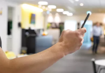 Person holder en mobiltelefon i sin hånd med et kontormiljø i baggrunden