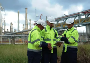 Tre arbetare i skyddsutrustning står framför ett olje- och gasraffinaderi, vilket symboliserar säkerheten för kritisk infrastruktur och skyddet av arbetskraften inom energibranschen.
