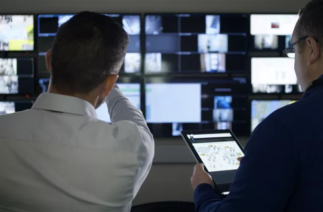 To personer står med ryggen til foran en væg fyldt med skærme med videoovervågningsbilleder på