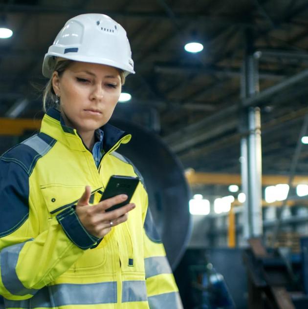 Female Industrial Worker in the Hard Hat Uses Mobile Phone While Walking Through Heavy Industry Manufacturing Factory. In the Background Various Metalwork Project Parts Lying