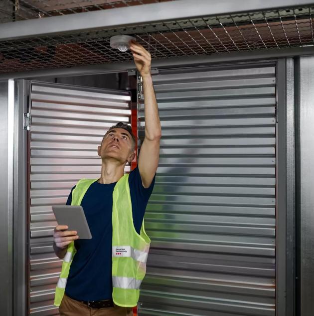 Fire alarm. Serious focused responsible man with tablet checking fire safety adjusting device on garage ceiling