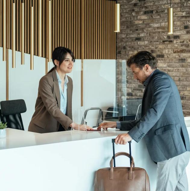 Traveling During COVID-19: Businessman with Luggage Passing Passport and Talking with Concierge on a Hotel Reception with Sneeze Guard Protection