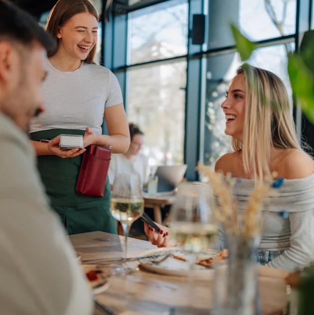 Smiling waitress holding pos terminal accepting payment from happy customers in modern restaurant
