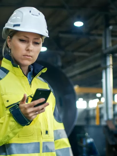Female Industrial Worker in the Hard Hat Uses Mobile Phone While Walking Through Heavy Industry Manufacturing Factory. In the Background Various Metalwork Project Parts Lying