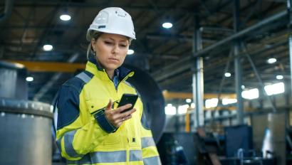 Female Industrial Worker in the Hard Hat Uses Mobile Phone While Walking Through Heavy Industry Manufacturing Factory. In the Background Various Metalwork Project Parts Lying