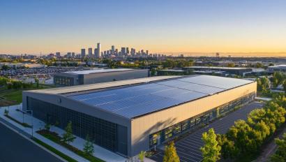 Aerial view of modern data center facility exterior, with solar panels on the roof and green space around it. The building is large in size, featuring sleek grey metal cladding and glass windows that showcase its interior tech equipment. In front of the structure stands an empty parking lot surrounded by trees, while the background shows other buildings and the city skyline under clear blue skies during golden hour. --ar 3:2 --quality 2 --v 6.1 Job ID: dcb08744-a35a-47be-bf95-20bc2b7a7922