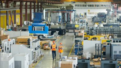 a man and a woman, both in orange vests and orange hard hats walk side-by-side through a warehouse