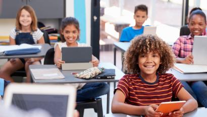 young children with smiles on their faces sit at their desks with tablets in front of them