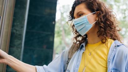 a woman wearing a yellow shirt and light blue jacket, with a mask on her face, is opening a door