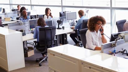 People in business attire sit in an open space workplace, talking on phones and working on their computers.