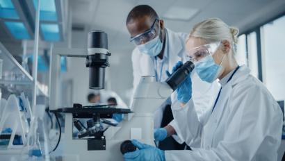 Modern Medical Research Laboratory: Two Scientists Wearing Face Masks Working Together Using Microscope, Analysing Samples, Talking. Advanced Scientific Lab for Medicine, Biotechnology.